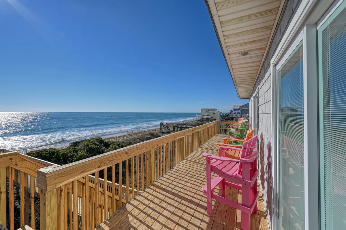 The image shows a spacious wooden deck overlooking the Atlantic Ocean, lined with vibrant Adirondack chairs. The sun is setting over the water, casting a warm glow across the deck—a perfect spot for relaxing, socializing, or simply soaking in the tranquil oceanfront ambiance.