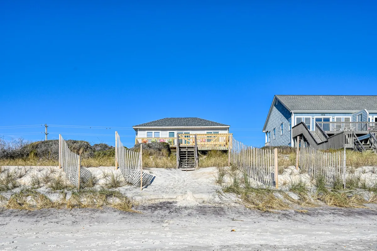 This photo depicts a classic beach house set against a backdrop of dunes and a sandy pathway leading directly to the beach. The fencing and open sky enhance the sense of privacy while keeping the beach within immediate reach.