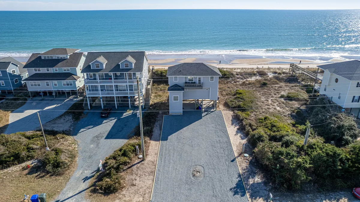 A wide-angle exterior shot of beachfront vacation homes with the ocean stretching out in the background. The sandy beach is clearly visible, and the area is depicted as uncrowded, ideal for sunbathing, beach walks, and family fun. The mood is relaxed and inviting, perfect for large groups and children building sandcastles.