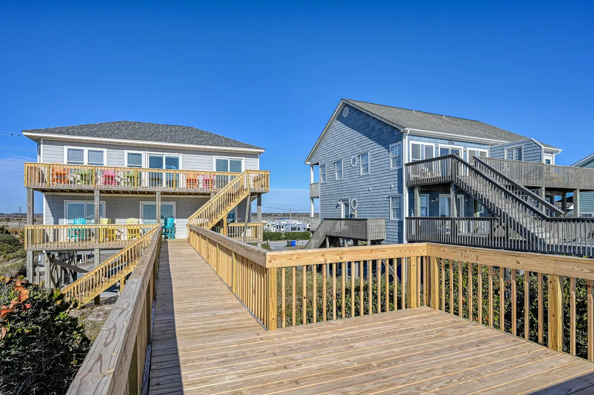 This image captures a sunlit exterior view with a private wooden boardwalk leading directly to the beach. The scene features colorful furniture scattered across wide balconies and two neighboring beach houses, evoking a sense of fun and togetherness. Families are seen relaxing and gathering on the decks, overlooking the ocean.