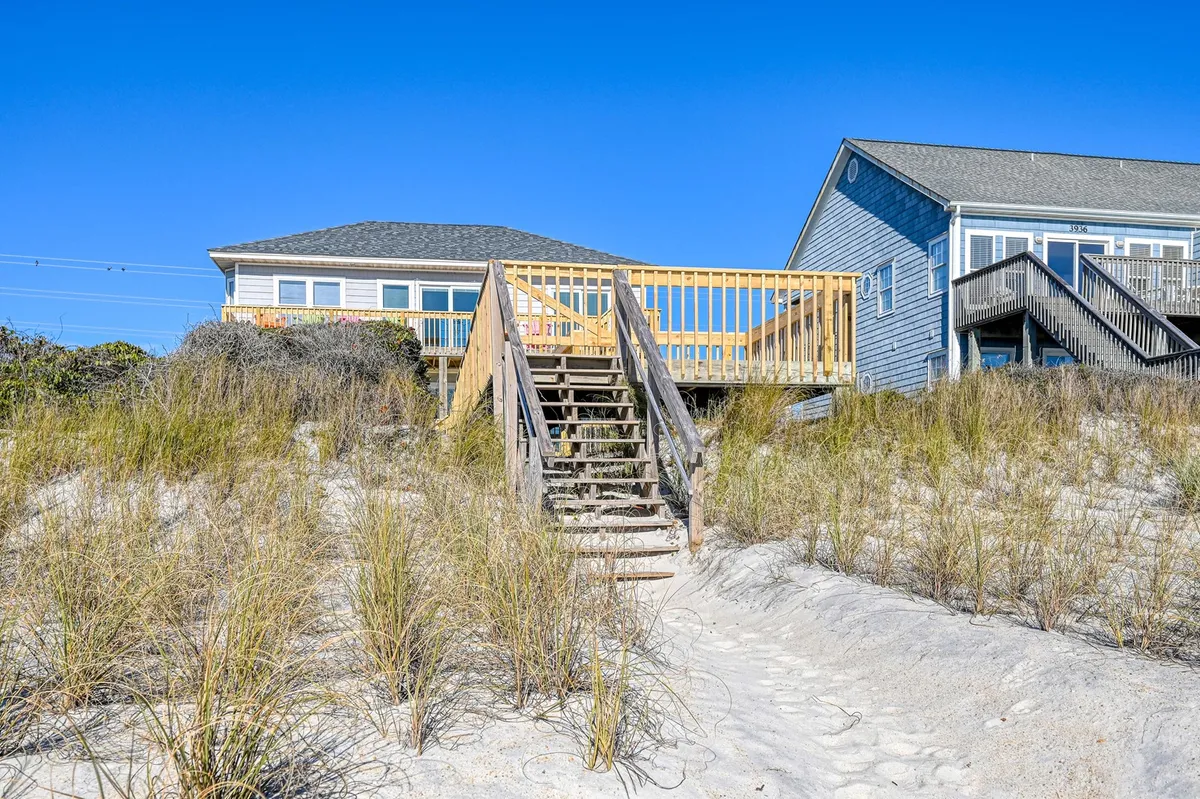 This exterior image shows the beach house with its inviting wooden steps leading up to a sunlit deck, where comfortable seating is arranged for enjoying ocean views. The sandy path in the background hints at direct access to the beach, and the overall mood is relaxed and welcoming, perfect for vacationers eager for outdoor adventures.