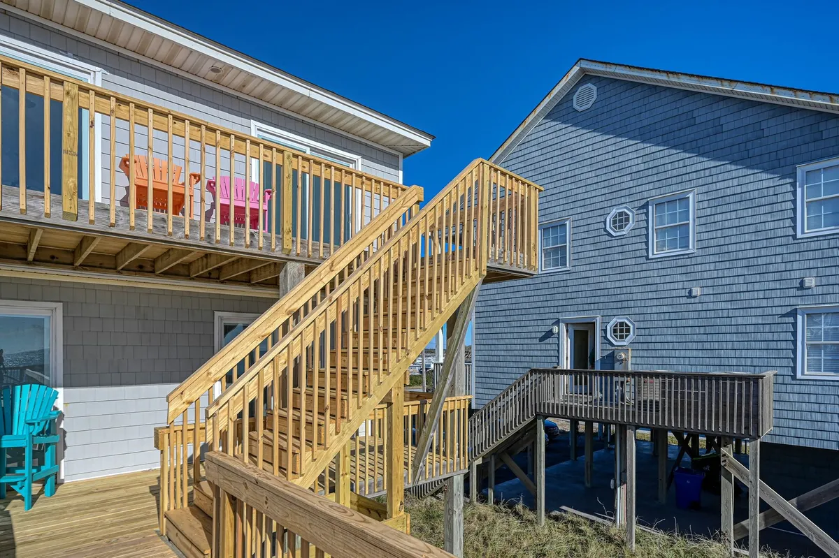 A cheerful exterior scene featuring a wooden porch with colorful chairs, the house's exterior walls, and a welcoming staircase. The deck is oriented to catch sunset views, and the setup invites socializing and enjoying quiet moments while overlooking the water or relaxing after a day of exploration.