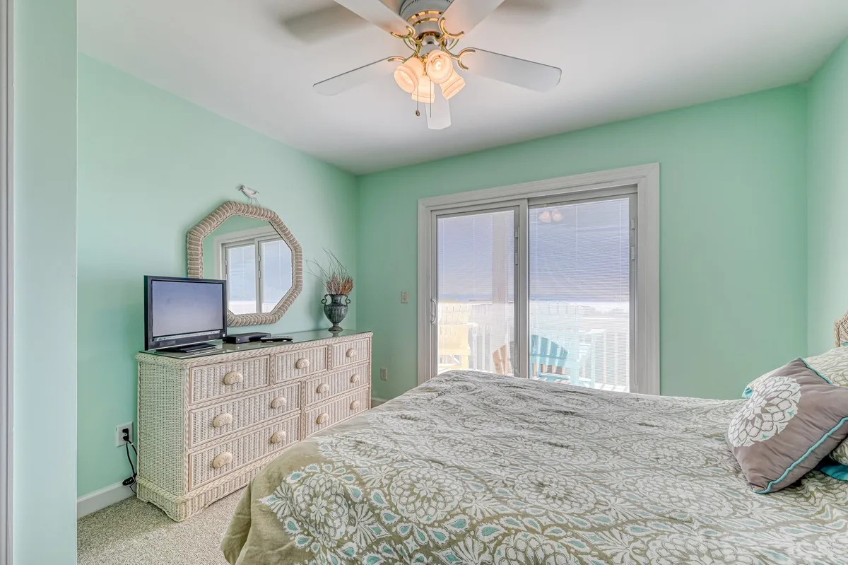 A cozy queen suite featuring a decorative bed, wicker dresser with mirror, and sliding glass doors leading to a balcony. The mood is serene and comfortable, and the view through the doors hints at the proximity to the ocean, ideal for private relaxation or watching sunsets.