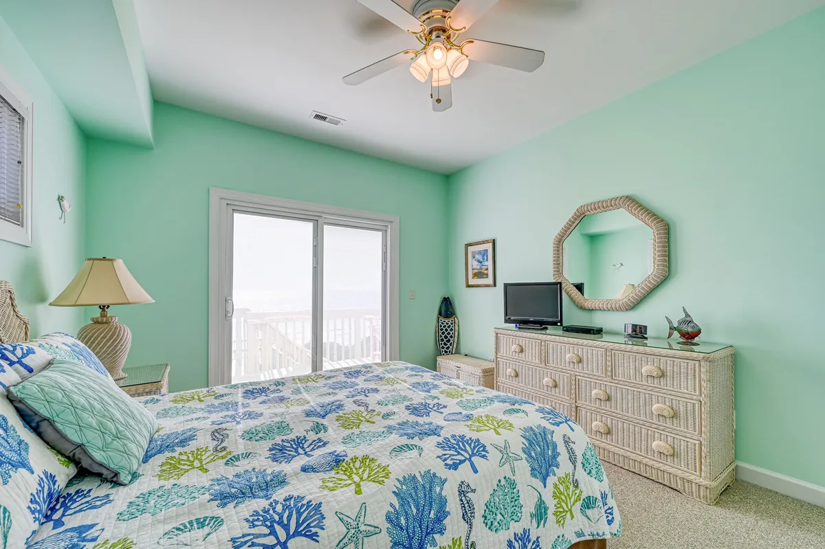 A queen bedroom with colorful bedding, a dresser and lamp, and a sliding glass door that leads directly to a private balcony. The room is filled with natural light and offers tranquil ocean views, creating a serene atmosphere perfect for relaxation after a day at the beach.