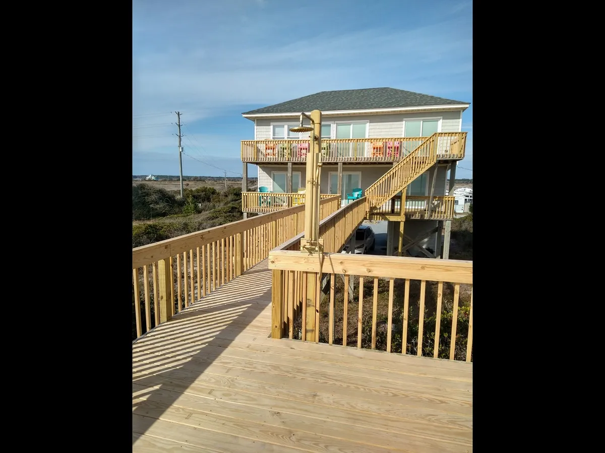 This image shows the welcoming exterior of the beach house, featuring a spacious wooden deck, a prominent staircase leading to the entry, and the house facade surrounded by natural landscape. The area appears inviting for gatherings, outdoor dining, and provides clear access from the parking area to the entry.