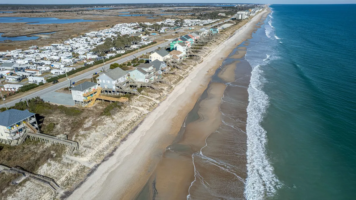 This image presents a sweeping view of beachfront houses lined along the coast, with gentle ocean waves, a scenic coastal road, and grassy dunes in the foreground. The setting is bright and relaxed, encapsulating the spirit of beachside living.